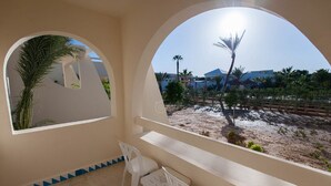 A room with a large arched window, a small table with two chairs, and a view of a sandy area with palm trees and buildings. - Cesar Thalasso (Djerba Midun)