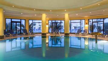 An indoor swimming pool with clear glass walls, blue lounge chairs, and a view of palm trees outside.