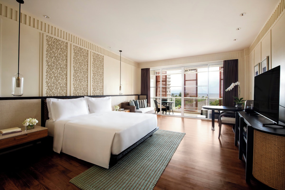 A hotel room with a bed, a desk with a TV, a balcony with a table and chairs, and a view of the outdoors.