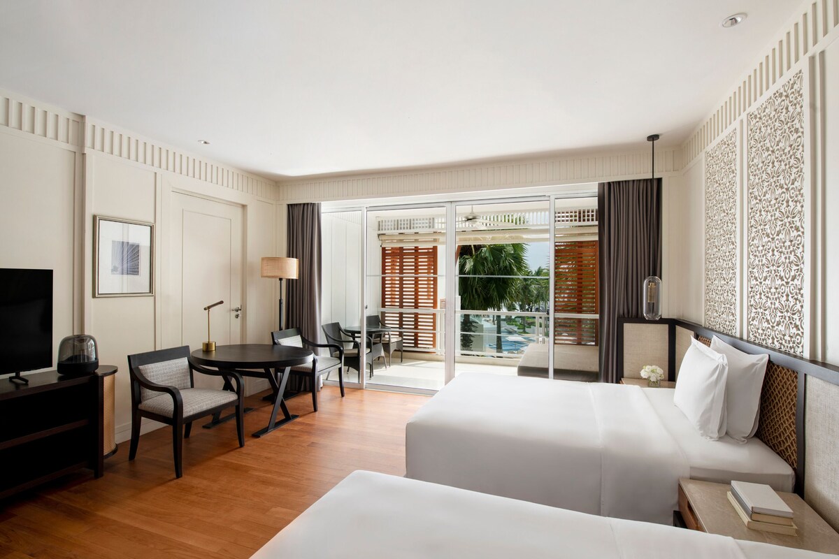 A hotel room with a bed, a desk with a chair, a TV, and a balcony with a view of the outdoors.