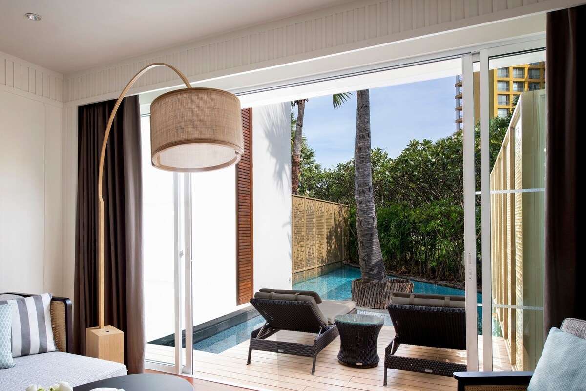 A modern hotel room with a large sliding glass door leading to a pool area, a sofa, a small table, and a floor lamp.