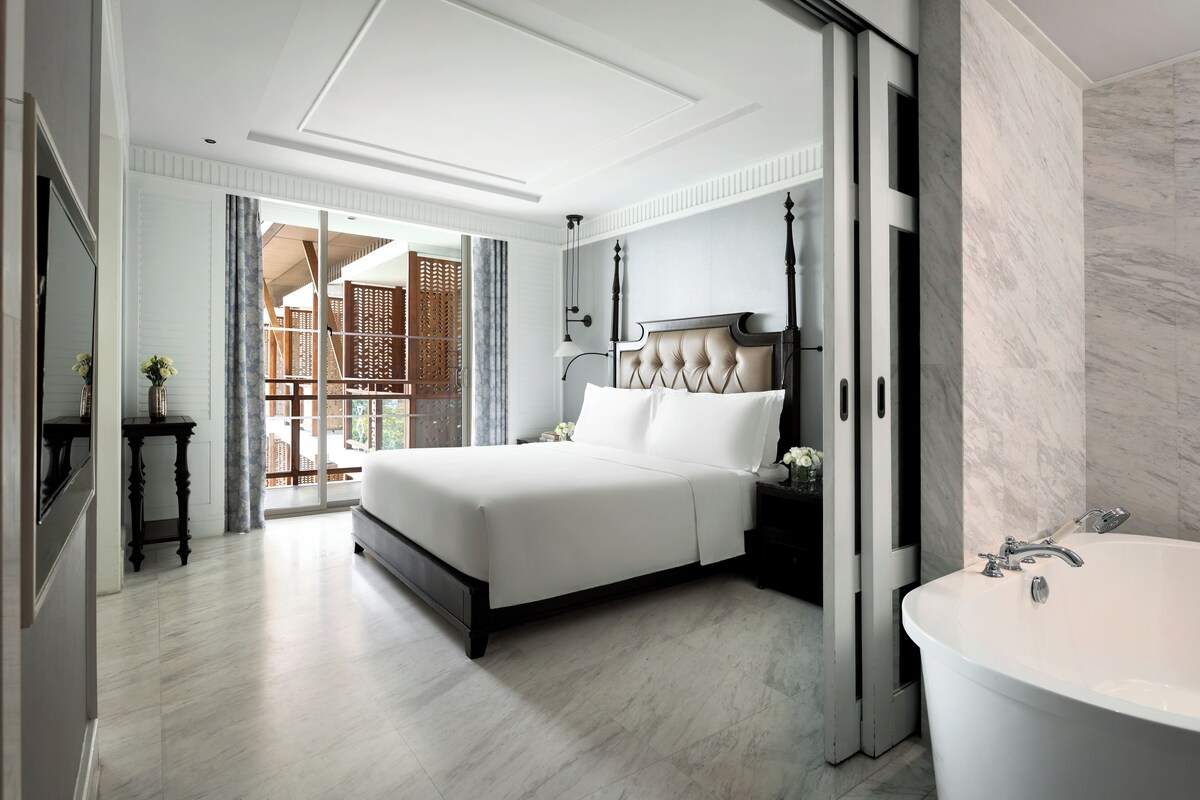 A modern bedroom with a large bed, a separate bathroom with a bathtub, and a view of a wooden balcony.