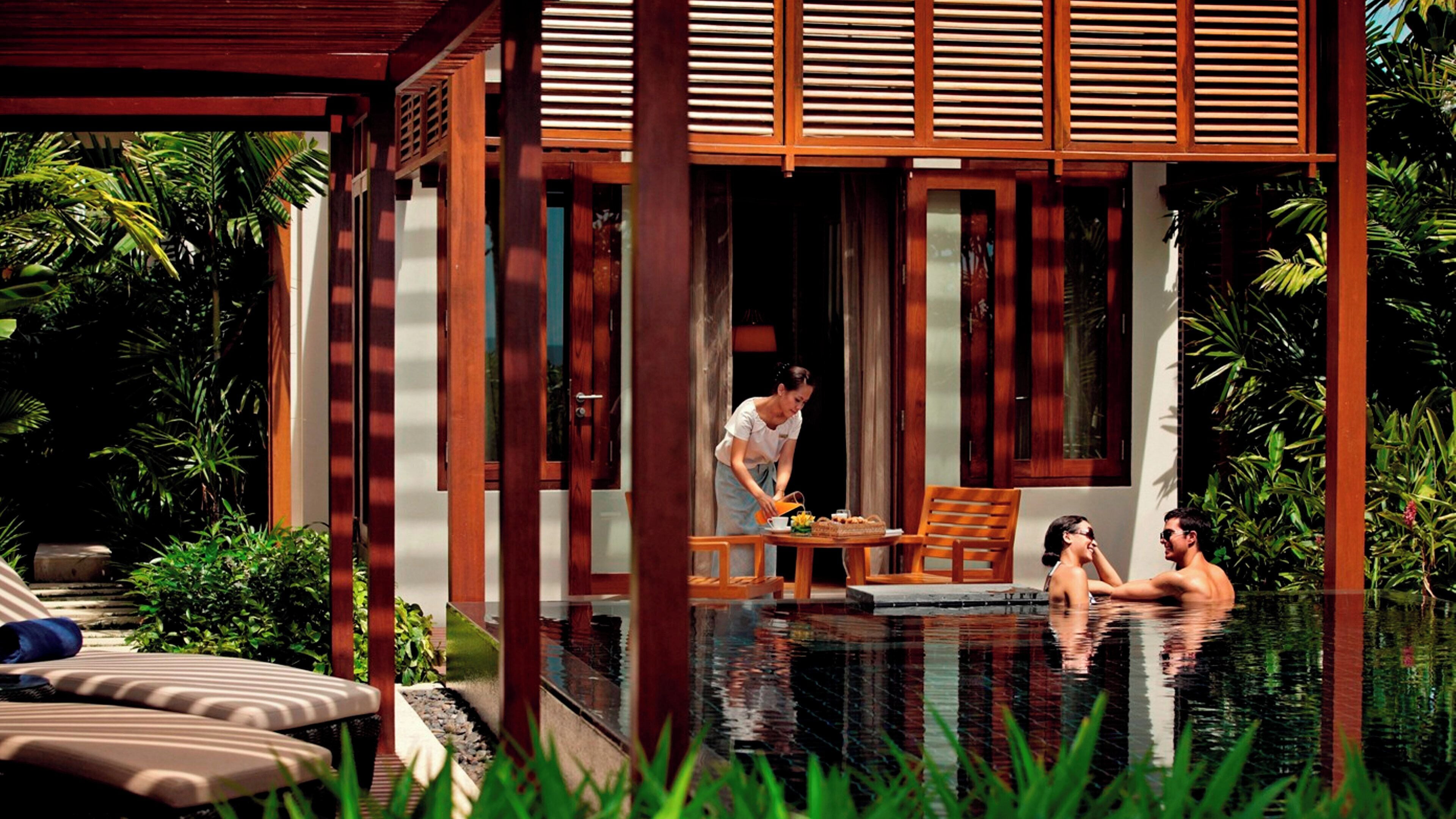 A poolside area with a wooden deck, lounge chairs, and a person tending to a table.