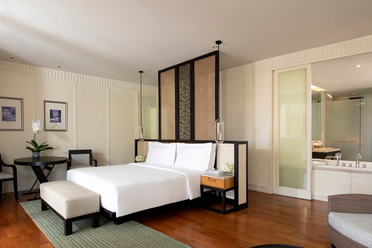A hotel room with a large bed, a desk with a chair, a small table with a vase of flowers, and a large mirror.
