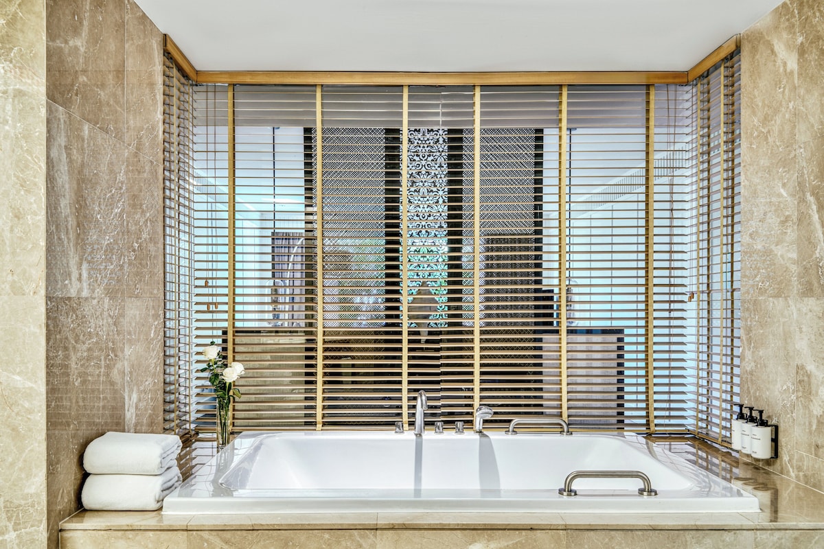 Un baño moderno con una bañera grande, paredes de mármol y una ventana decorativa con persianas.
