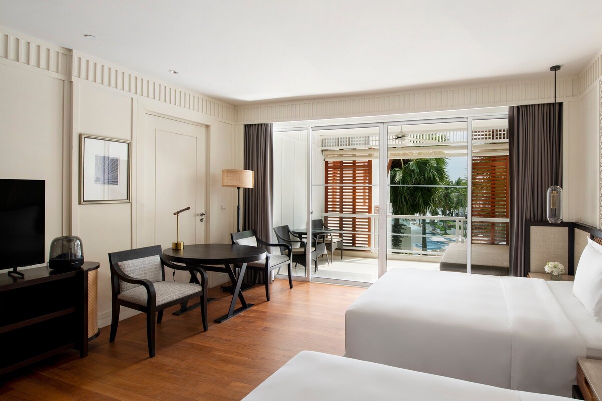 A hotel room with a bed, a desk with a chair, a TV, a lamp, and a view of a pool and palm trees through the glass door.