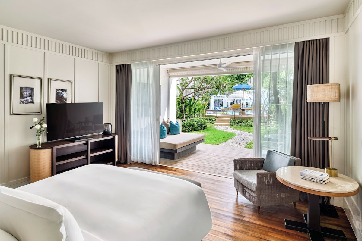Habitación de hotel con una cama grande, televisión, una mesita de noche con lámpara y vistas a una piscina exterior.