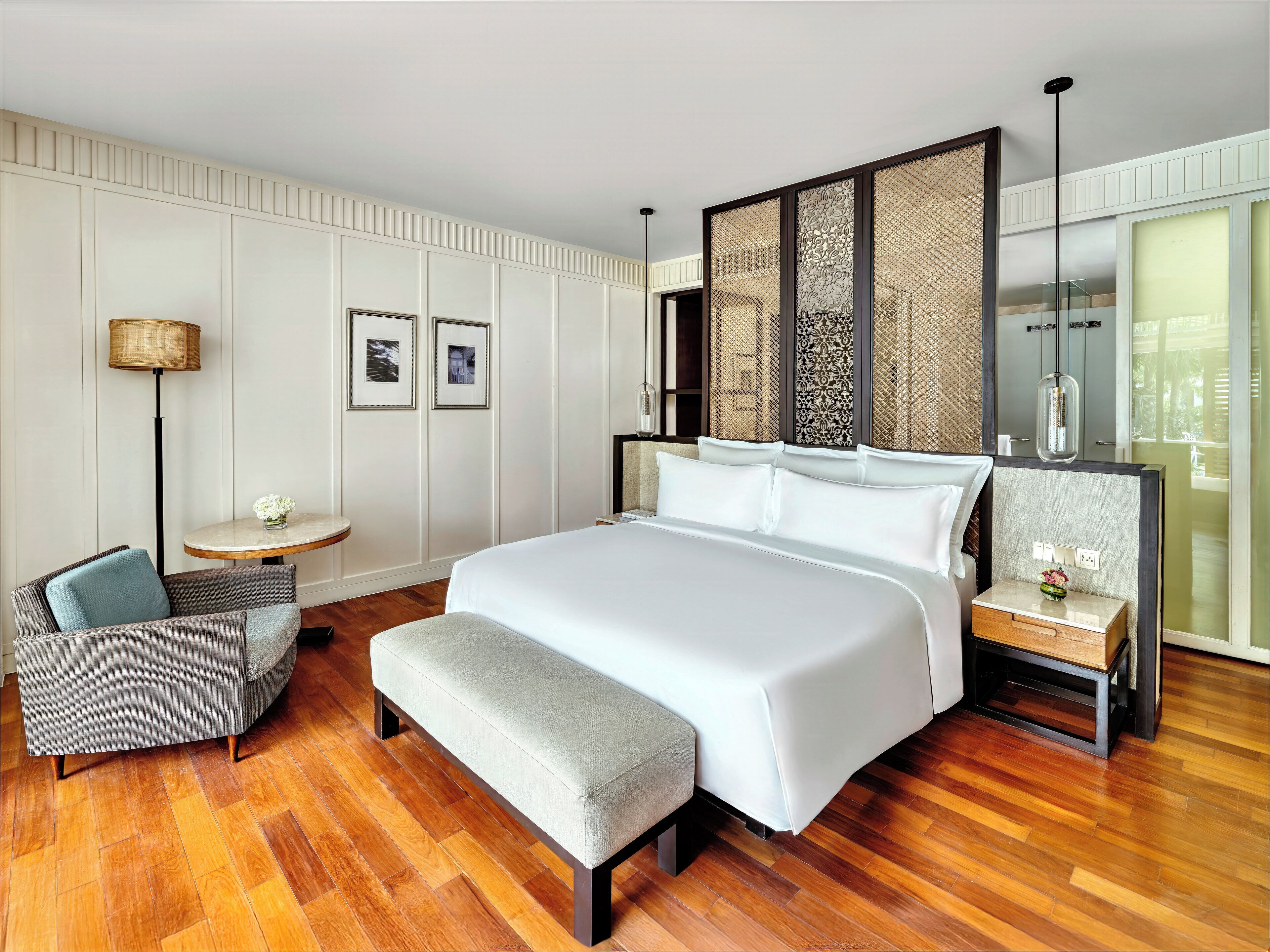 Une chambre d’hôtel moderne dotée d’un grand lit, d’un coin salon avec un fauteuil et une petite table, d’un panneau décoratif mural et d’une grande fenêtre.