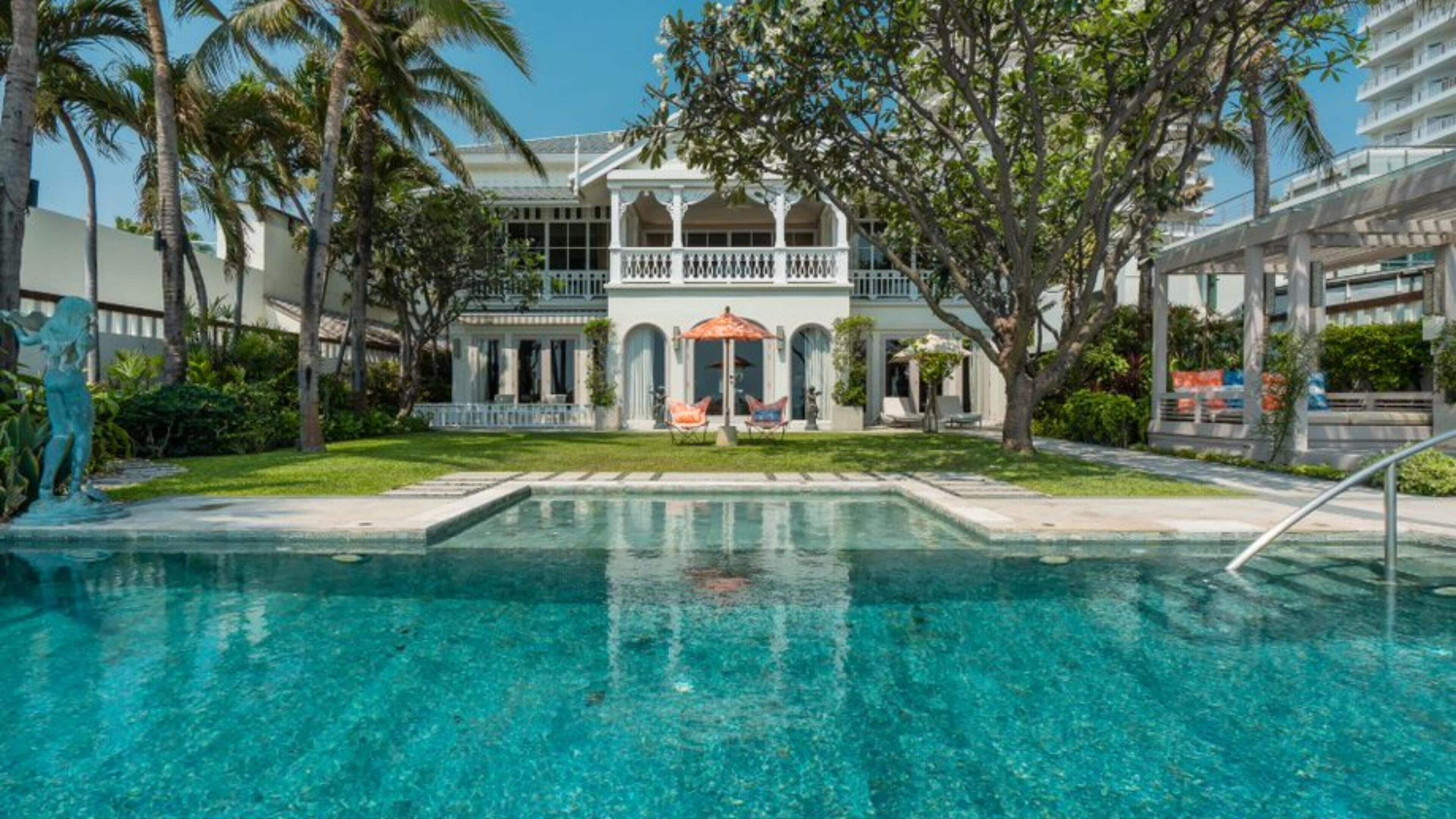 Una piscina con superficie de agua azul cristalina, rodeada de palmeras y un edificio de fachada blanca y ventanas de arco.