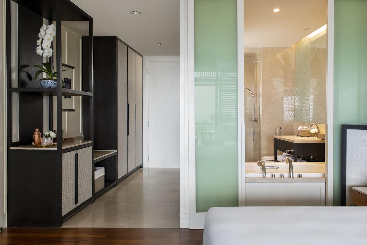 Une chambre moderne avec un grand lit, une armoire sombre, une salle de bain dotée d’une douche à parois de verre et un petit coin salon agrémenté d’une plante.