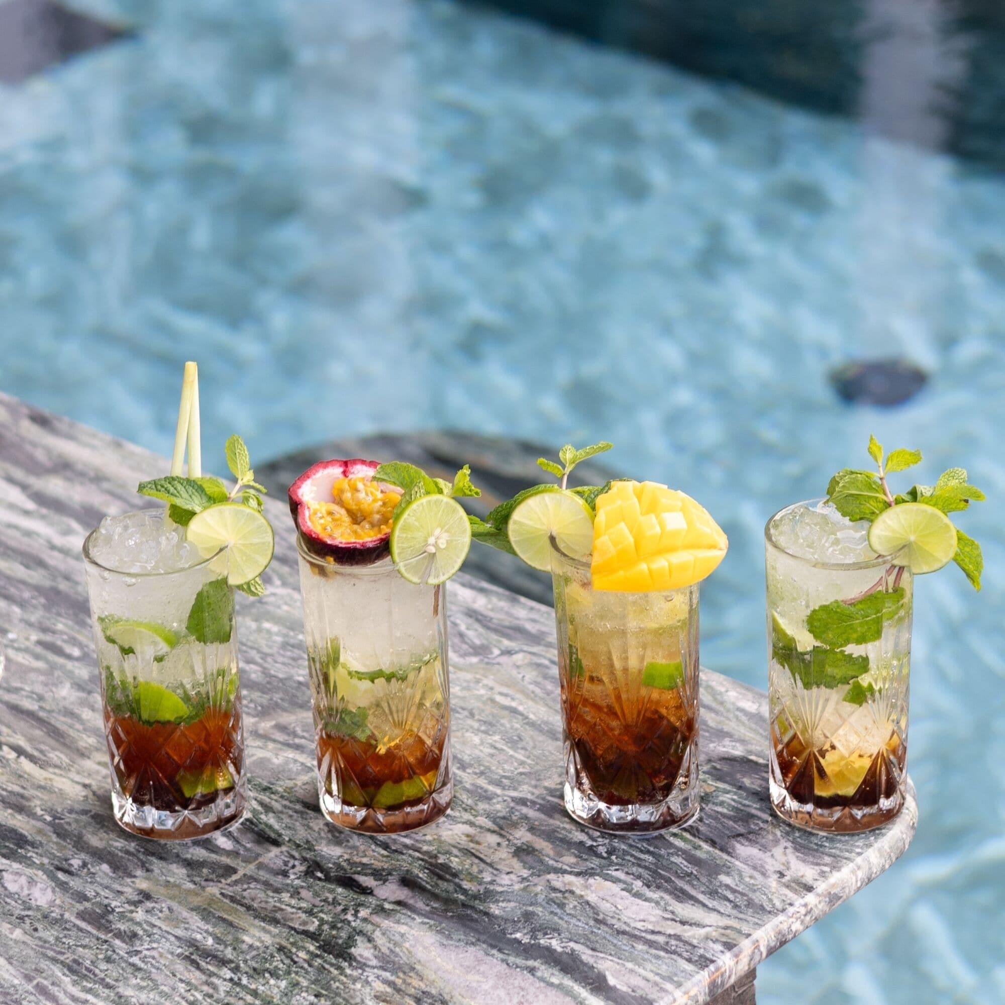 Cuatro vasos de bebidas heladas con diversas frutas y hojas de menta, sobre una superficie de mármol cerca de una piscina.