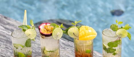 Cuatro vasos de bebidas heladas con diversas frutas y hojas de menta, sobre una superficie de mármol cerca de una piscina.