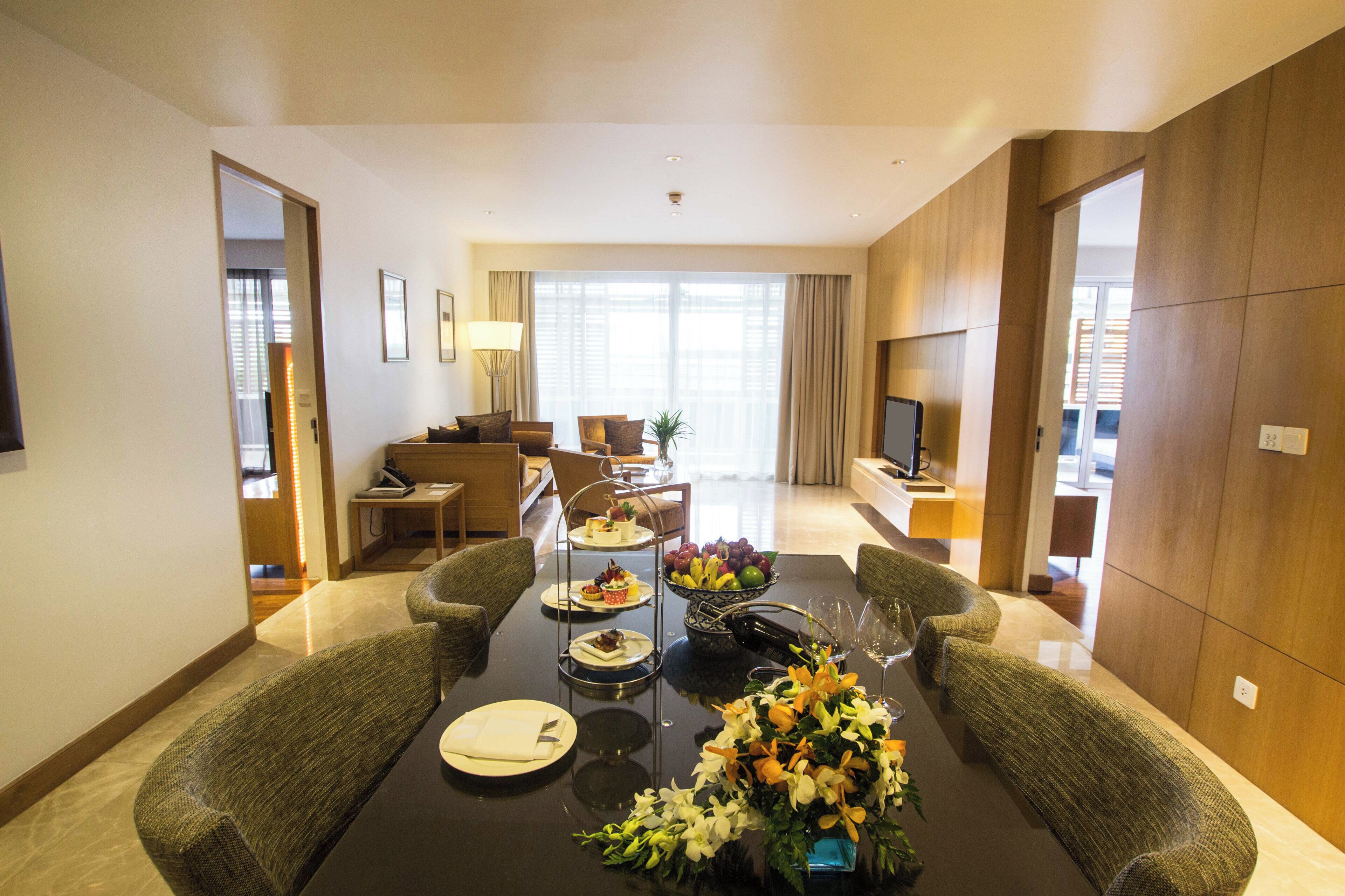 A hotel room with a dining table set for four, a sofa, a TV, and a large window.