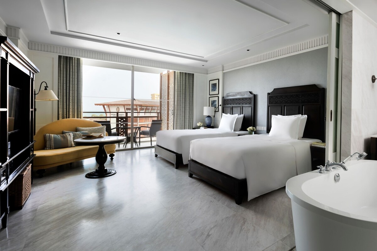 A hotel room with two beds, a sitting area with a sofa and table, a large bathtub, and a balcony with a view.