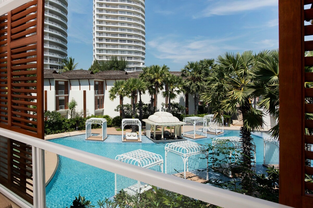 A pool area with multiple lounge chairs and umbrellas, surrounded by palm trees and a view of tall buildings.