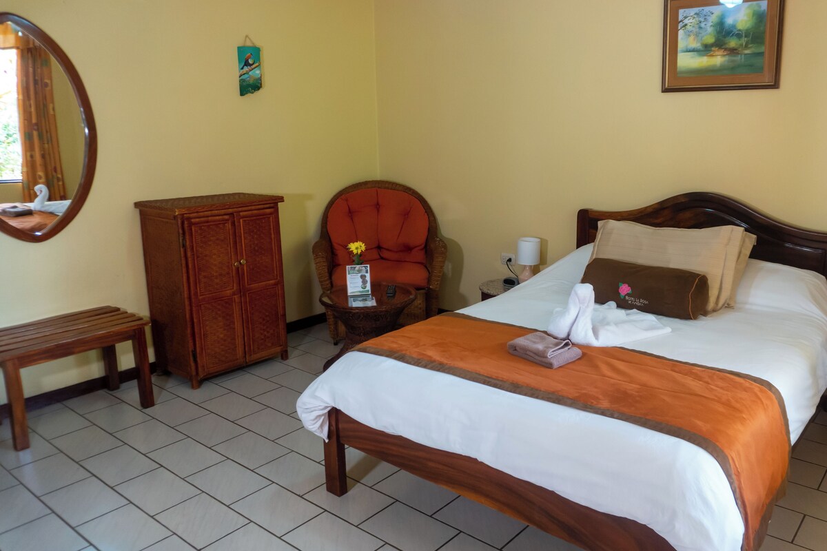 A hotel room with a bed, a wooden cabinet, a chair, a small table, a nightstand, and a wall-mounted picture.