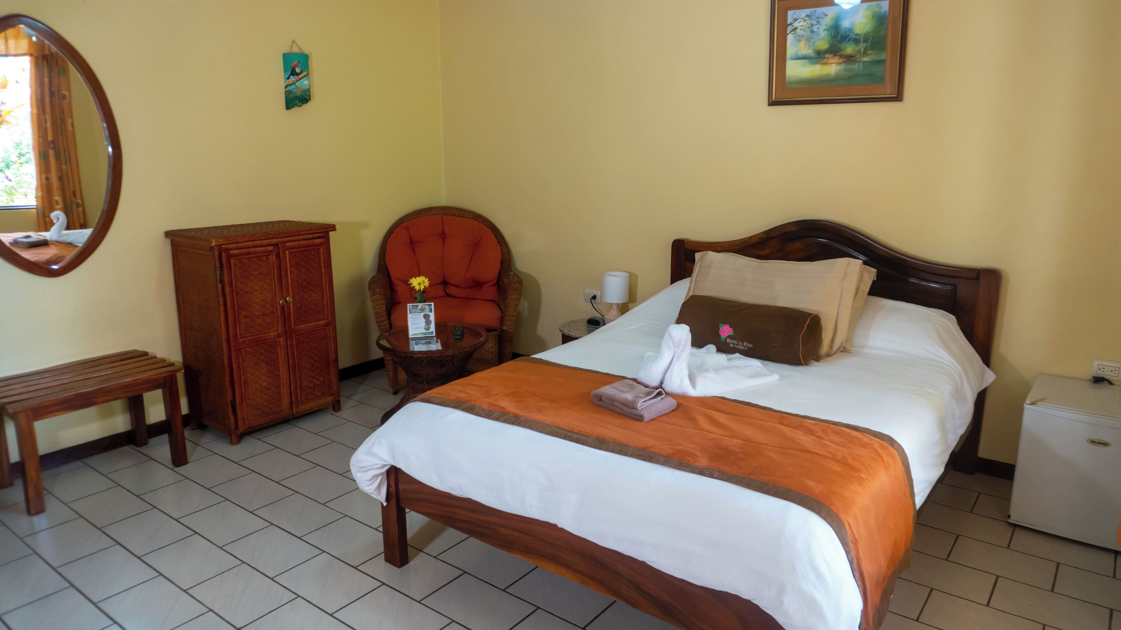 A hotel room with a bed, a wooden cabinet, a chair, a small table, a nightstand, and a wall-mounted picture.