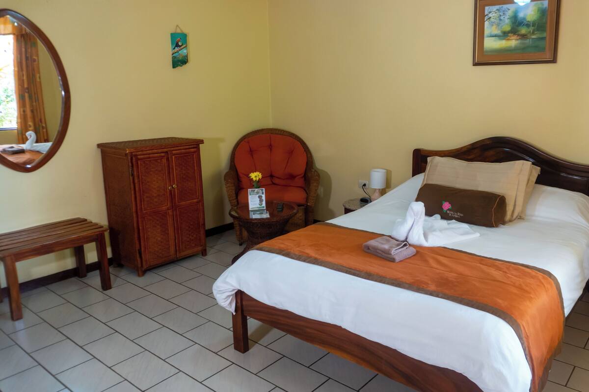 A hotel room with a bed, a wooden cabinet, a chair, a small table, a nightstand, and a wall-mounted picture.