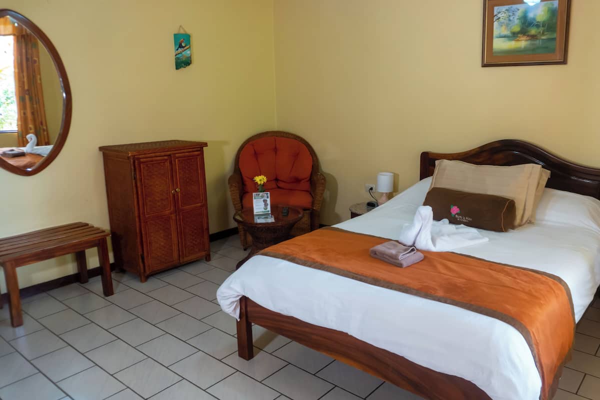 Ein Hotelzimmer mit Bett, einem Holzschrank, einem Sessel, einem kleinen Tisch, einem Nachttisch und einem an der Wand hängenden Bild.