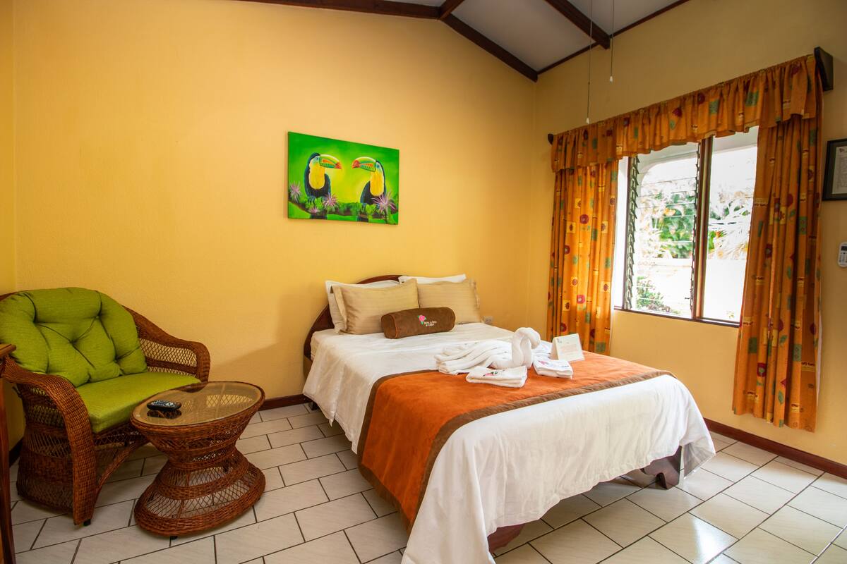 A hotel room with a bed, a green chair, a small table, and a painting of toucans on the wall.