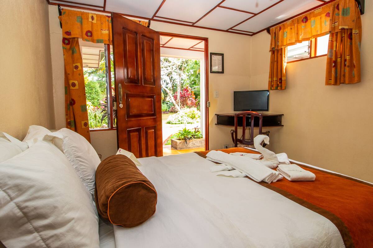 A hotel room with a bed, a television, and a view of the garden through an open door.