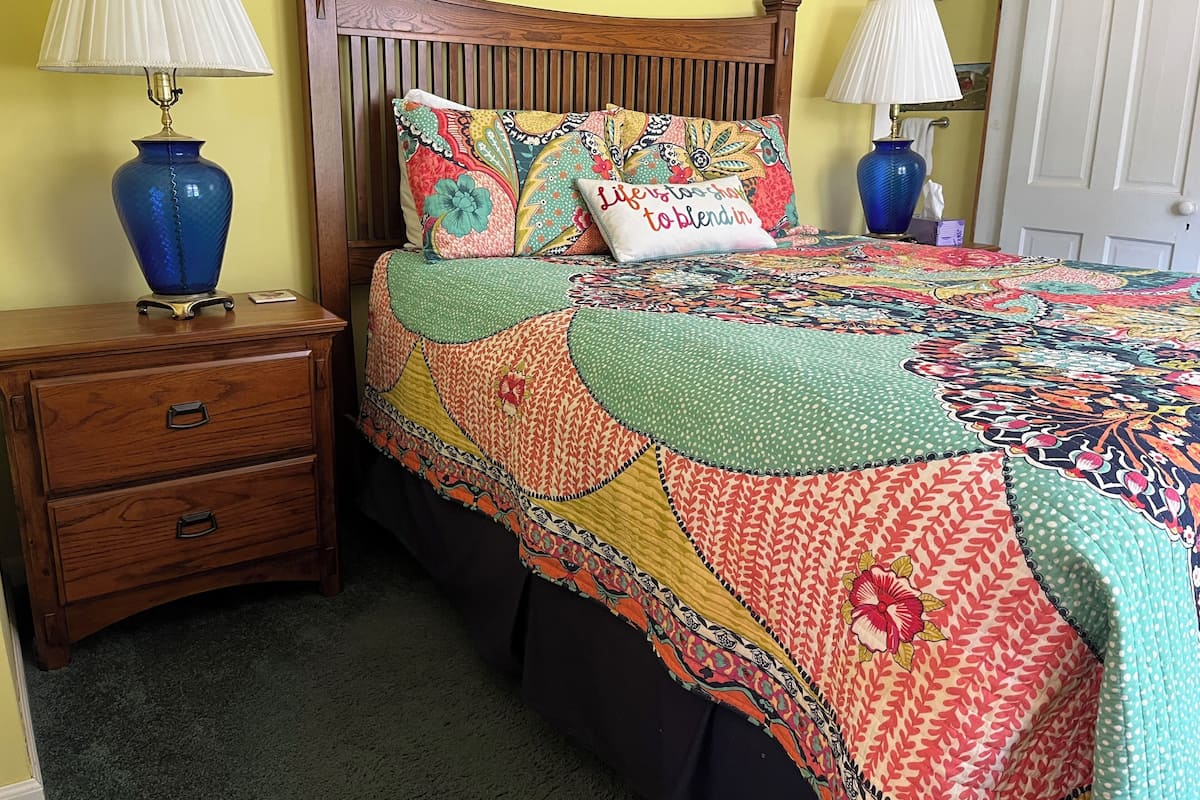 A bedroom with a wooden bed, a colorful quilt, two lamps, and a wooden nightstand with drawers.