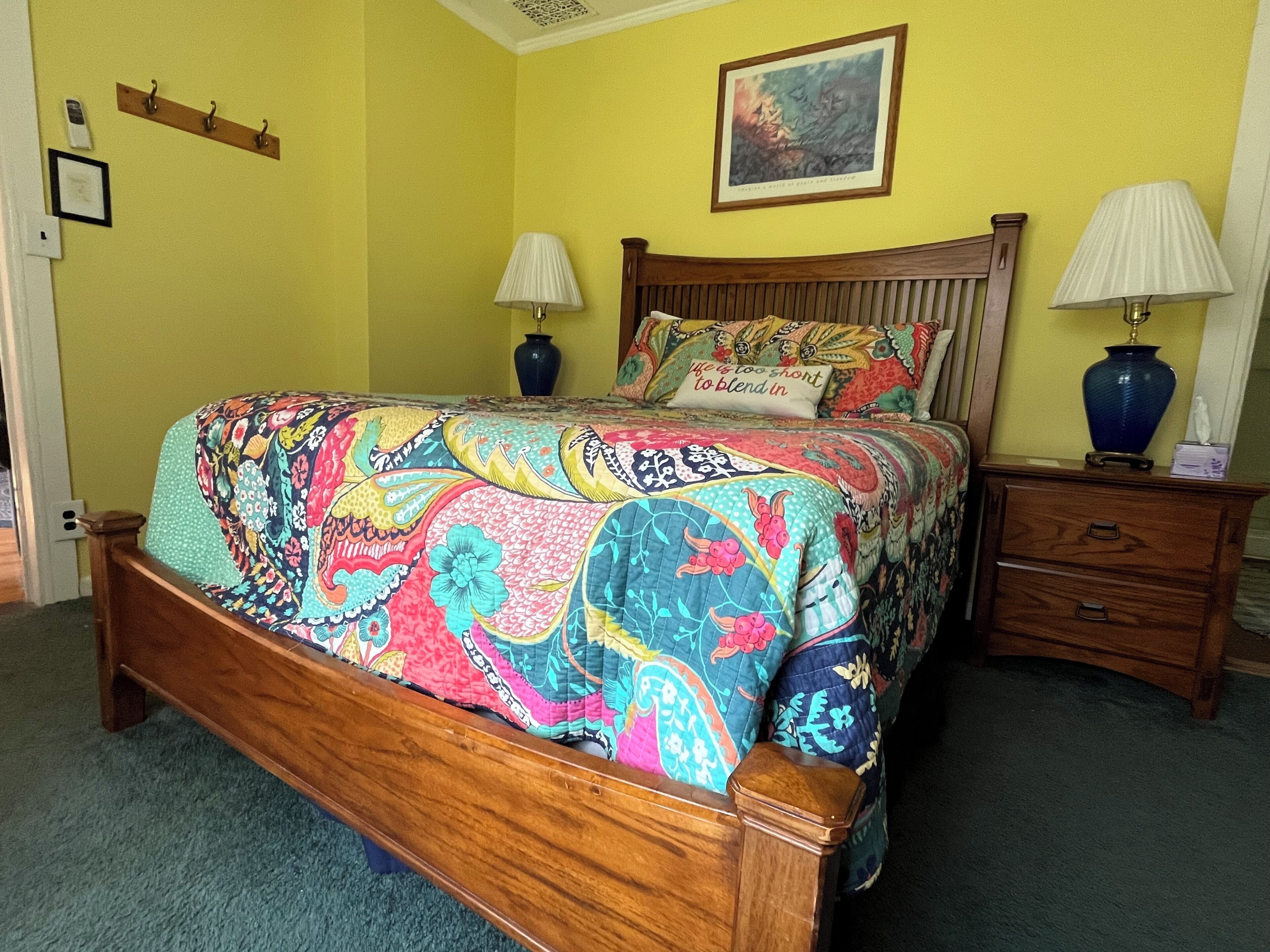 A bed with a colorful quilt, wooden headboard, two bedside tables with lamps, and a painting on the wall.