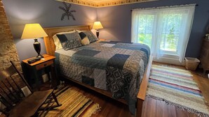 A bedroom with a bed, two bedside lamps, a nightstand, a chair, a window with curtains, and a decorative wall piece.