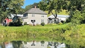 A two-story house with a porch, surrounded by lush greenery and a pond reflecting the building.