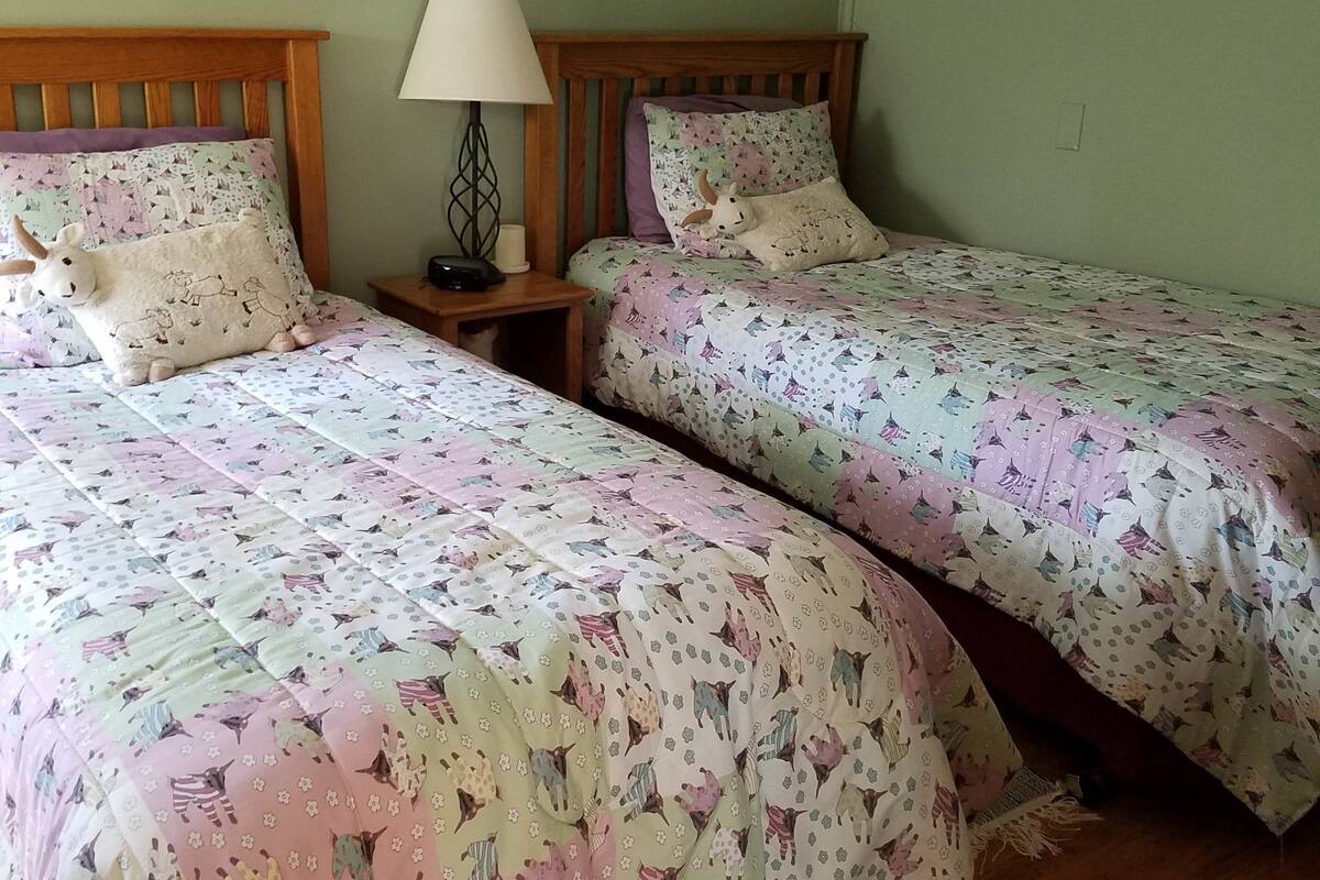 A room with two single beds, each with a floral-patterned bedspread and a wooden headboard. There is a nightstand with a lamp and a framed picture on the wall above the beds.
