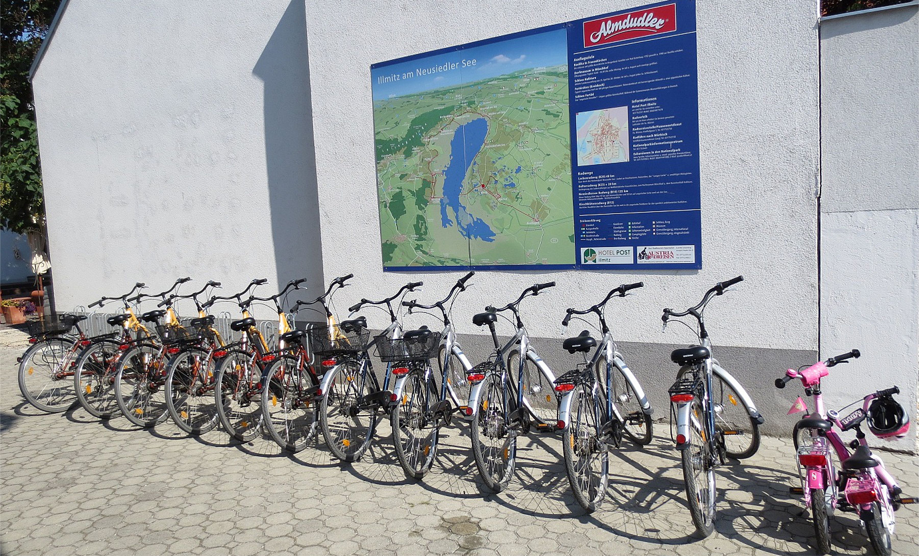 A row of bicycles parked against a building with a large map and information board on the wall.