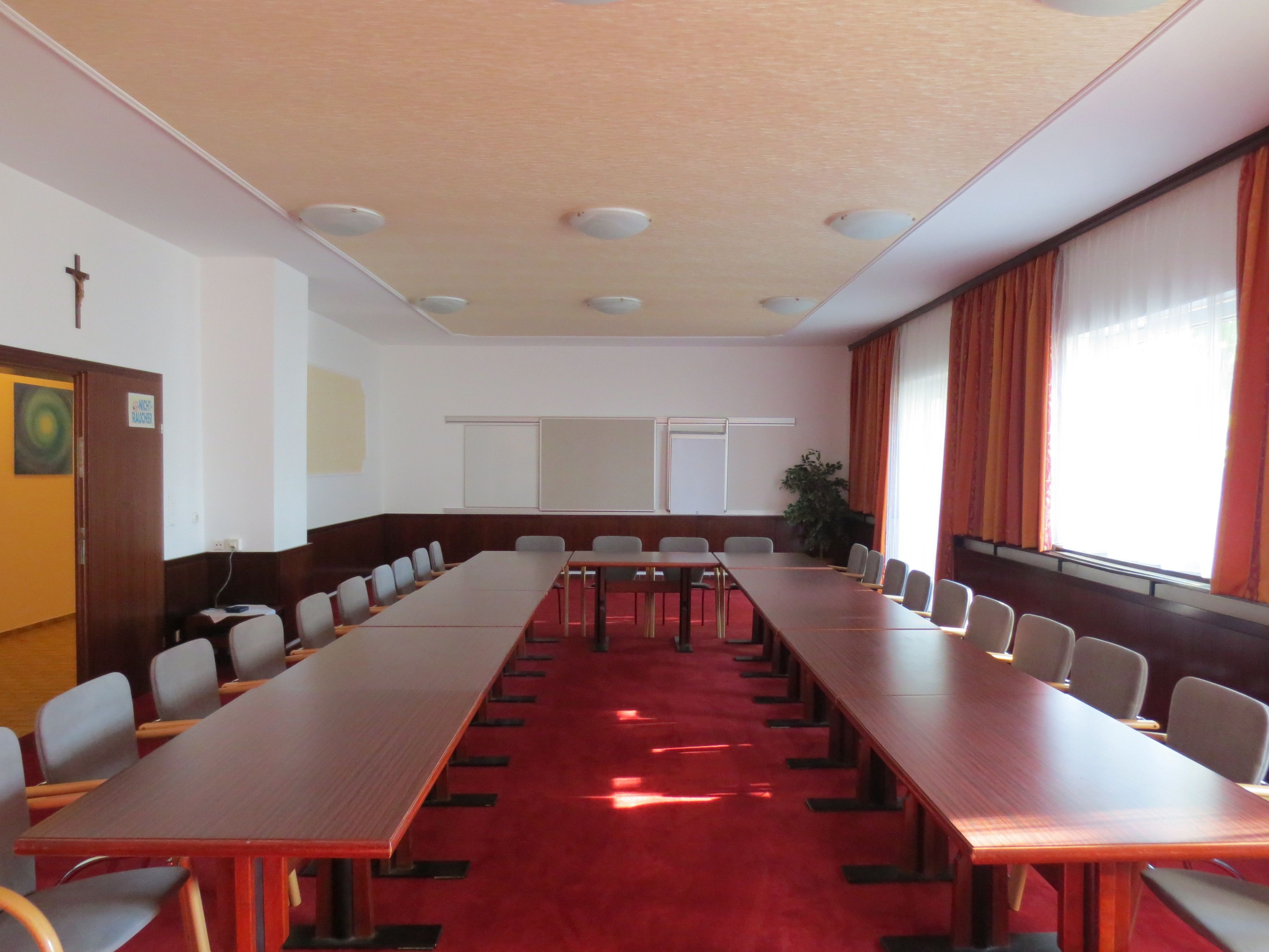 Une salle de conférence avec de longues tables, des chaises, un crucifix au mur et une plante.