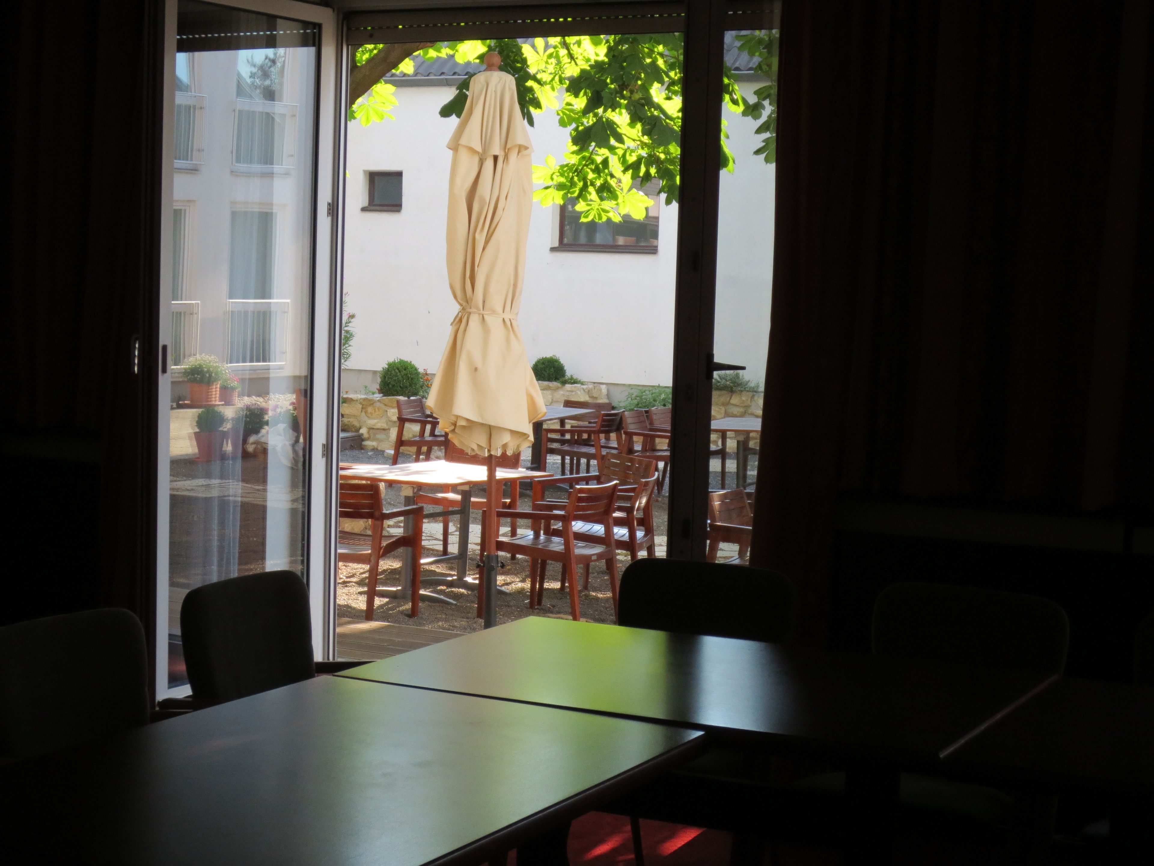 Ein Zimmer mit einer Schiebetür zu einer Terrasse im Freien mit Holztischen und -stühlen, einem beigen Sonnenschirm und Topfpflanzen.