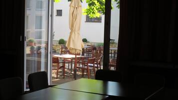 Ein Zimmer mit einer Schiebetür zu einer Terrasse im Freien mit Holztischen und -stühlen, einem beigen Sonnenschirm und Topfpflanzen.