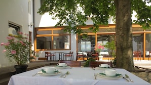 Un espace de restauration en plein air avec des tables pour deux personnes, entourées de chaises, et un grand arbre qui offre de l’ombre.