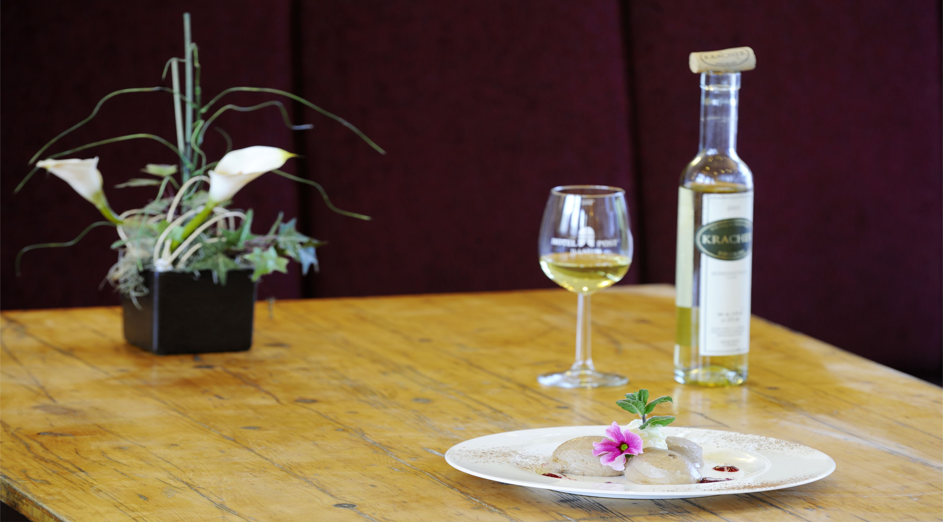 Een houten tafel met een bord gebakjes, een glas witte wijn en een fles Krächer wijn.
