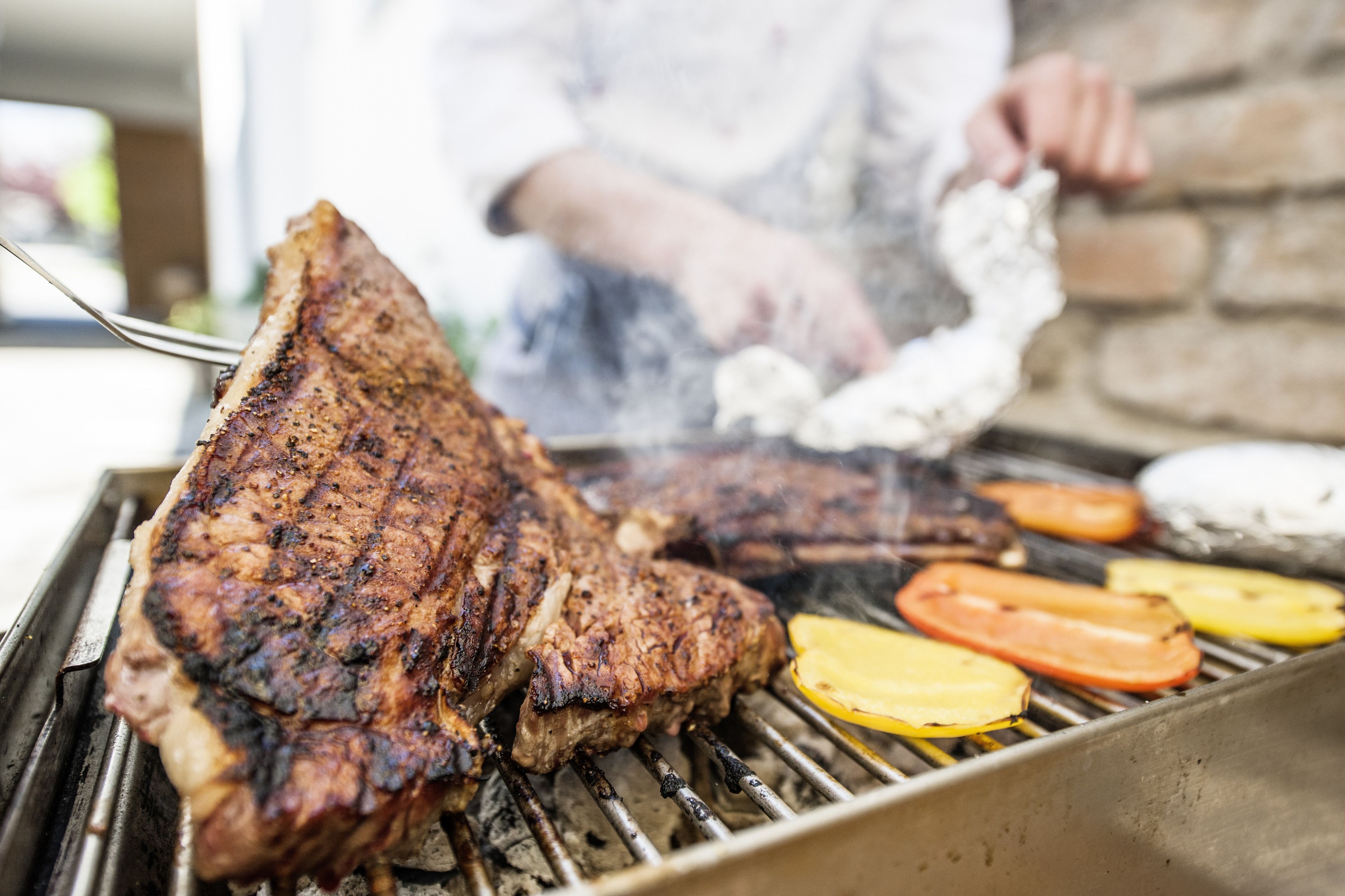 A steak being grilled on a barbecue with vegetables and a person in the background.