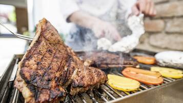 A steak being grilled on a barbecue with vegetables and a person in the background.