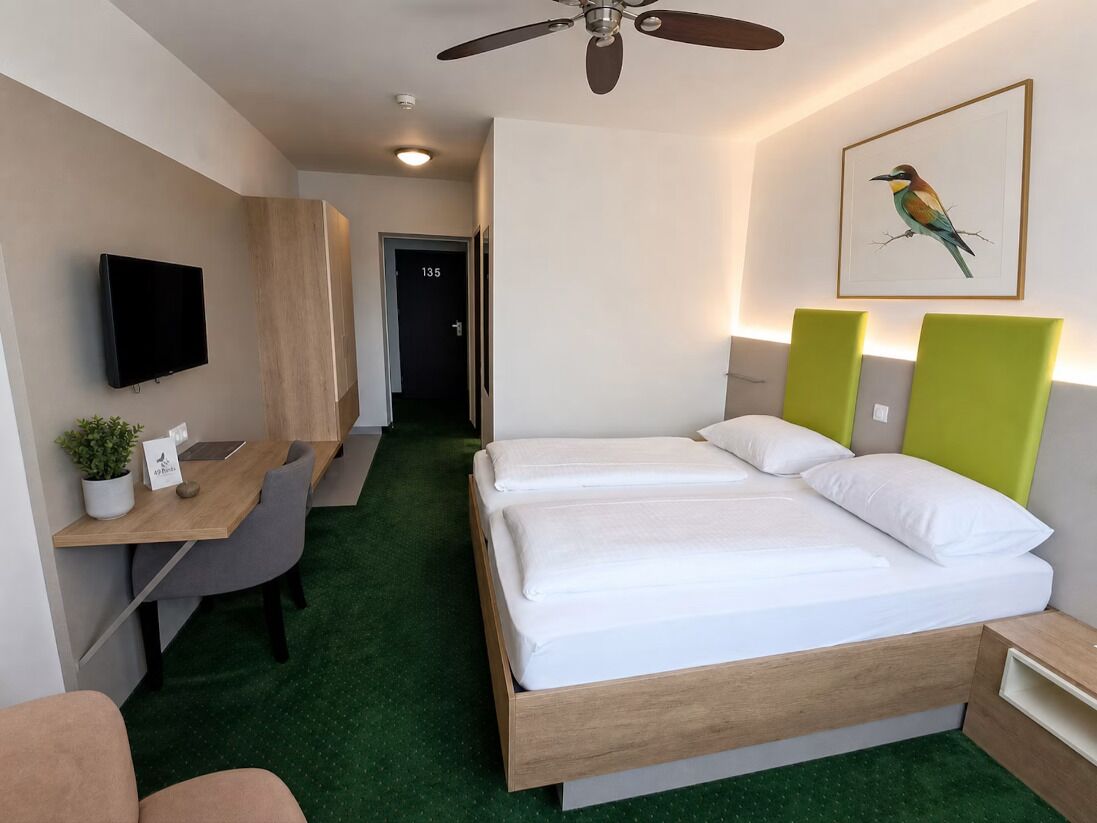 Quarto de hotel com cama, escrivaninha, cadeira, televisão e ventoinha no teto.