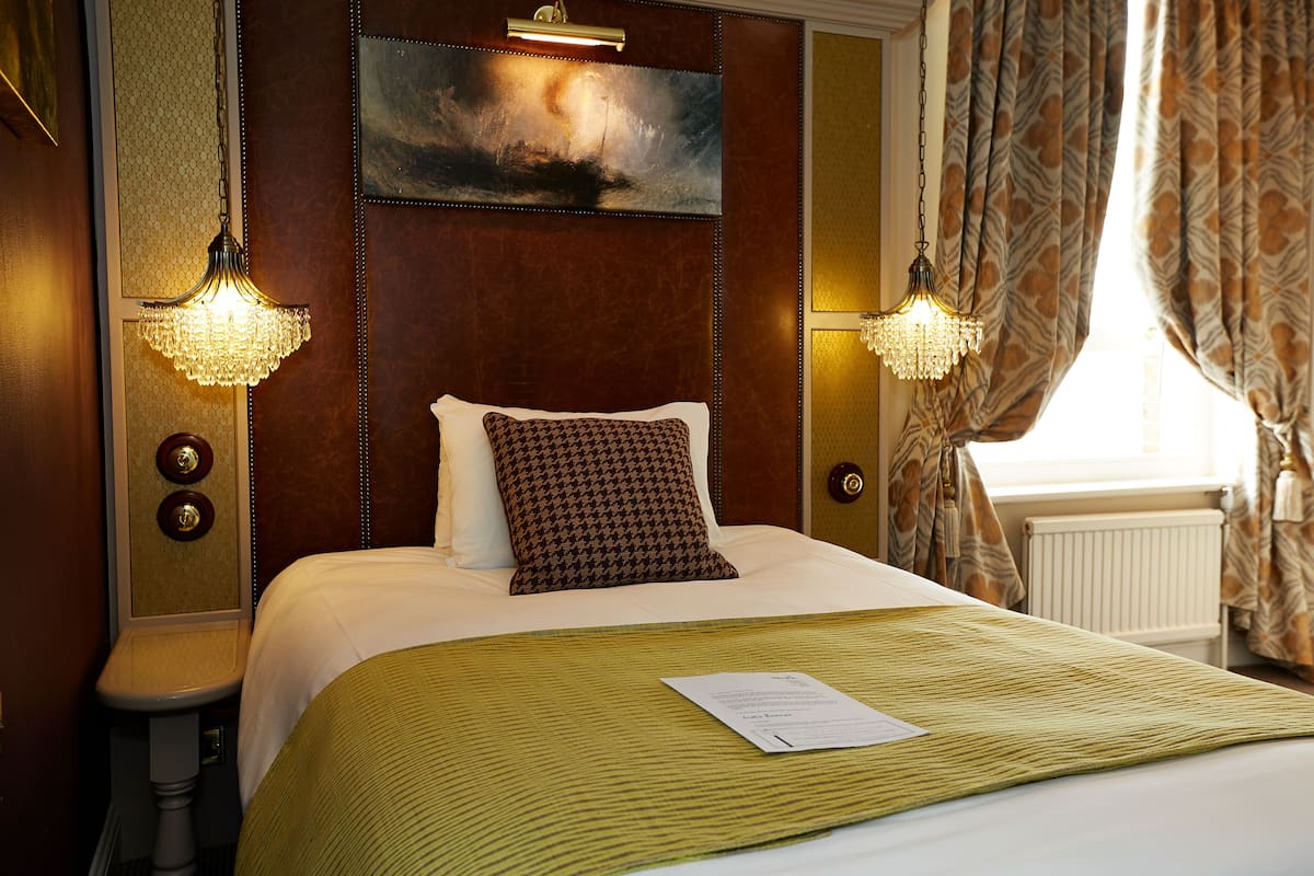 A hotel room with a large bed, two wall sconces, a painting above the bed, and a window with curtains.
