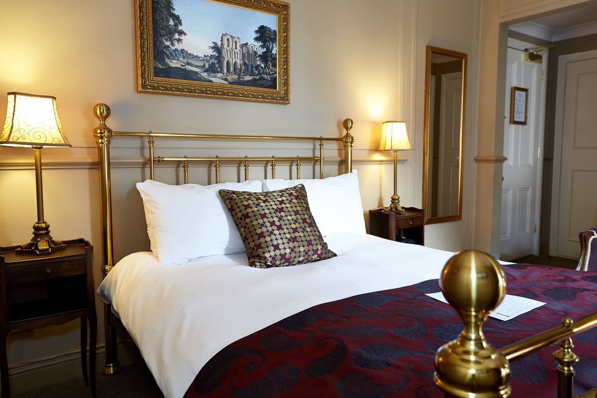 A neatly made bed with white and patterned pillows, a red and blue bedspread, and a gold-framed headboard. There are two bedside lamps on wooden nightstands.