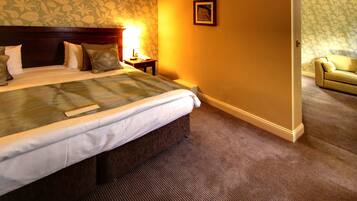 A hotel room with a large bed, a nightstand with a lamp, a framed picture on the wall, and a sofa in the adjacent room.