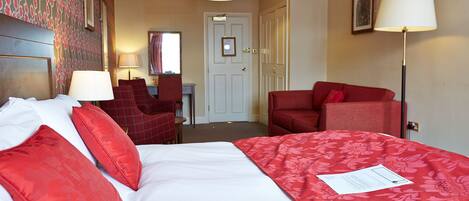 A hotel room with a large bed, red and white bedding, a red armchair, a desk with a chair, and a lamp.