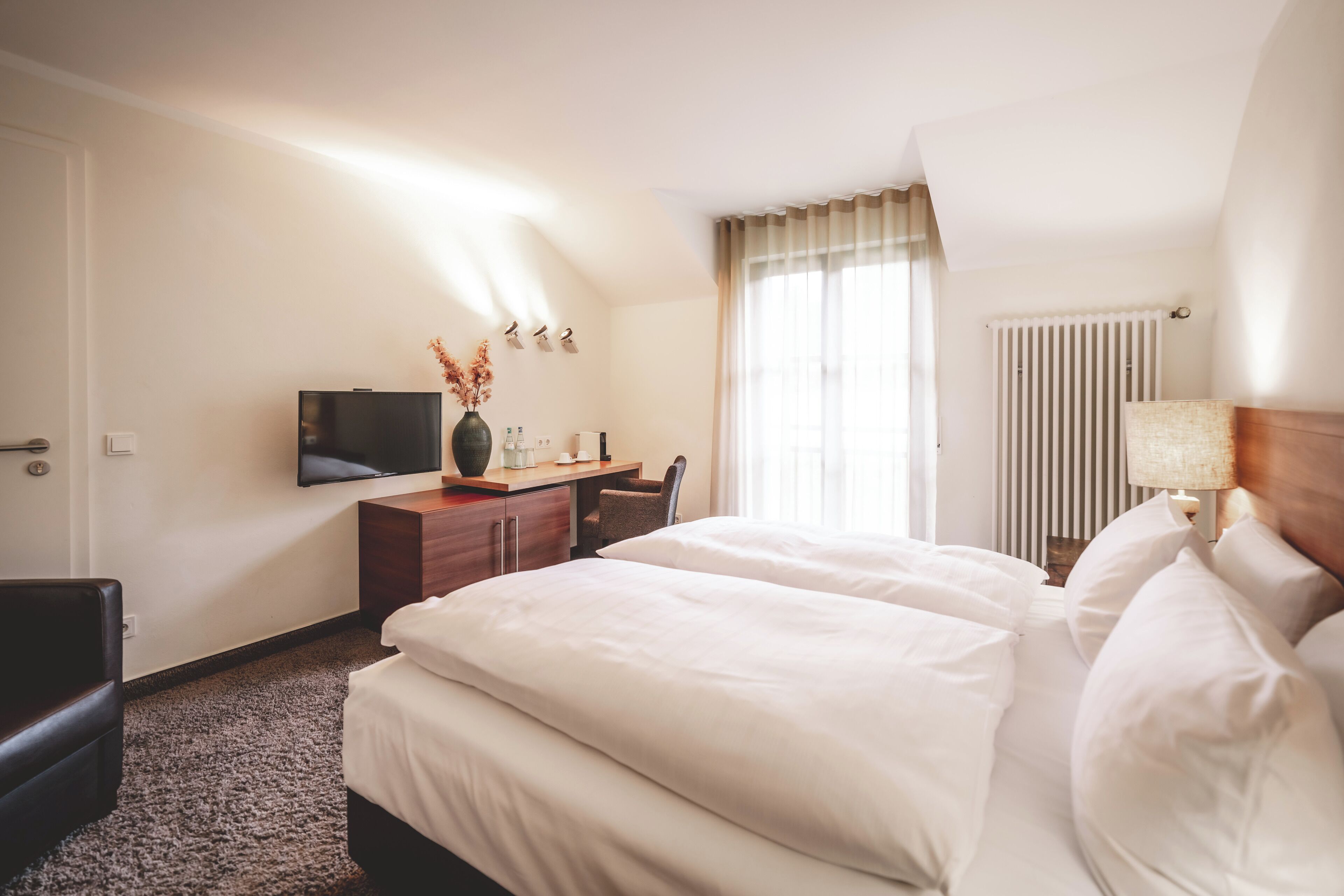 A hotel room with a large bed, a desk with a TV, a chair, and a radiator.