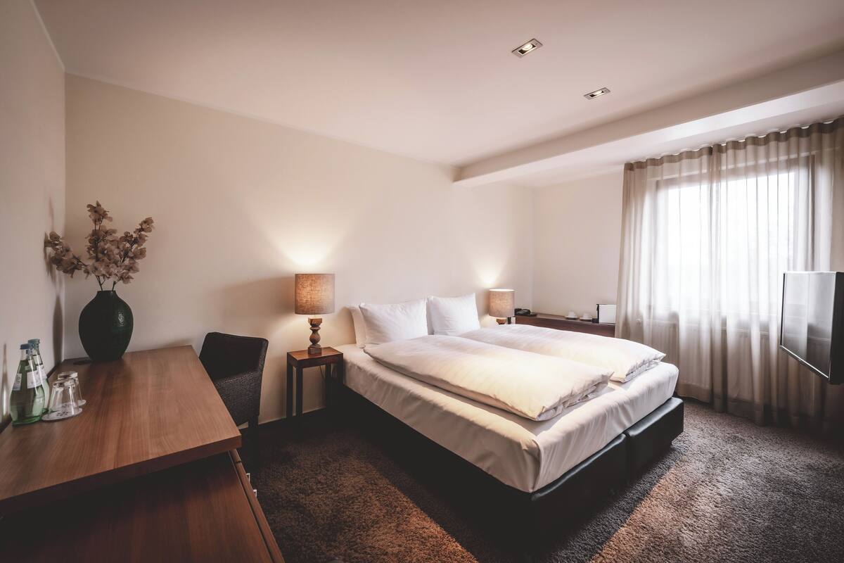 A hotel room with a large bed, a wooden desk, a vase with dried flowers, and a television.