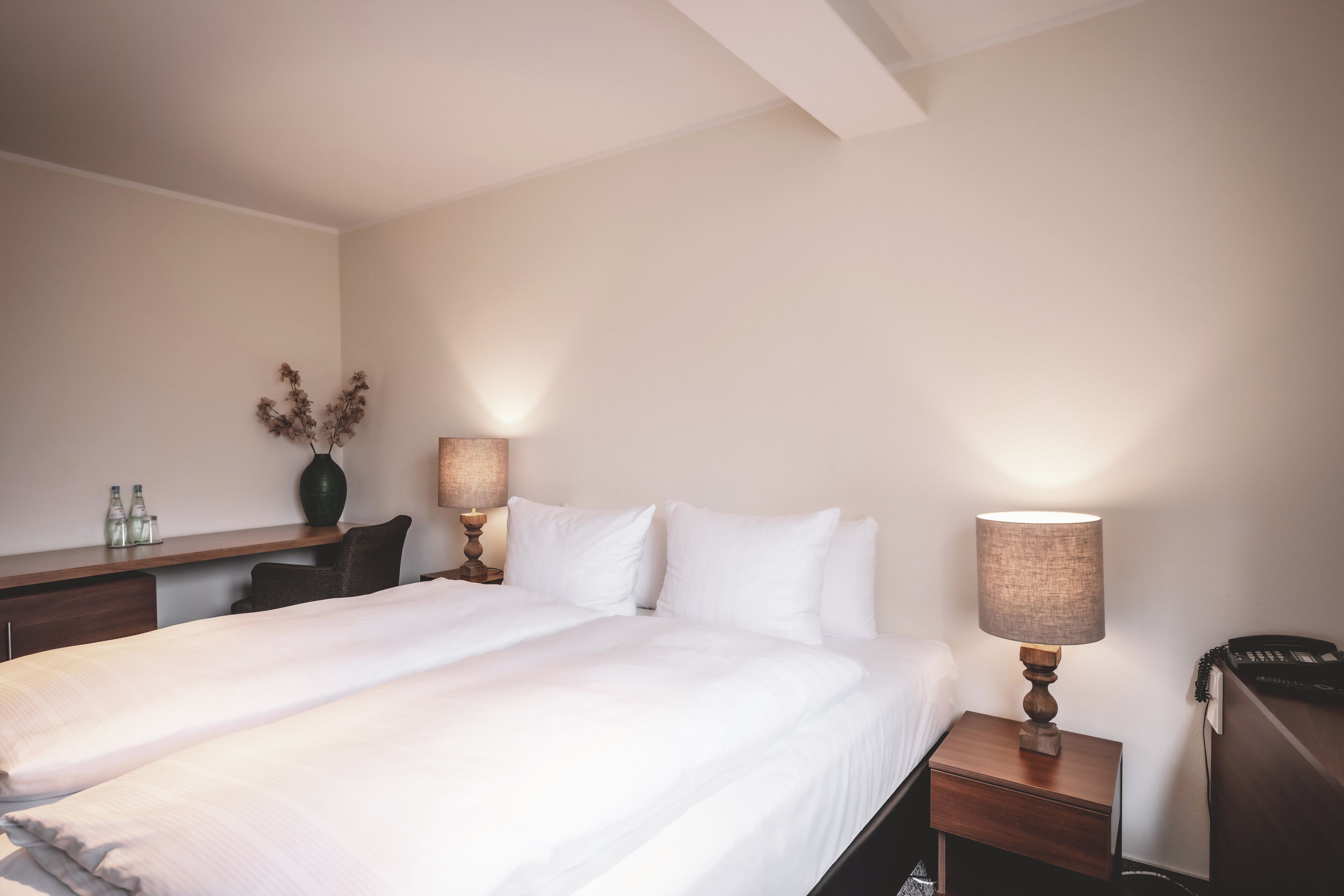 A hotel room with a large bed, a bedside table with a lamp, a desk with a vase and bottles, and a telephone.