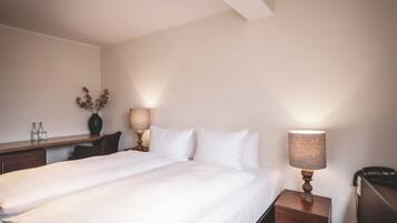 A hotel room with a large bed, a bedside table with a lamp, a desk with a vase and bottles, and a telephone.