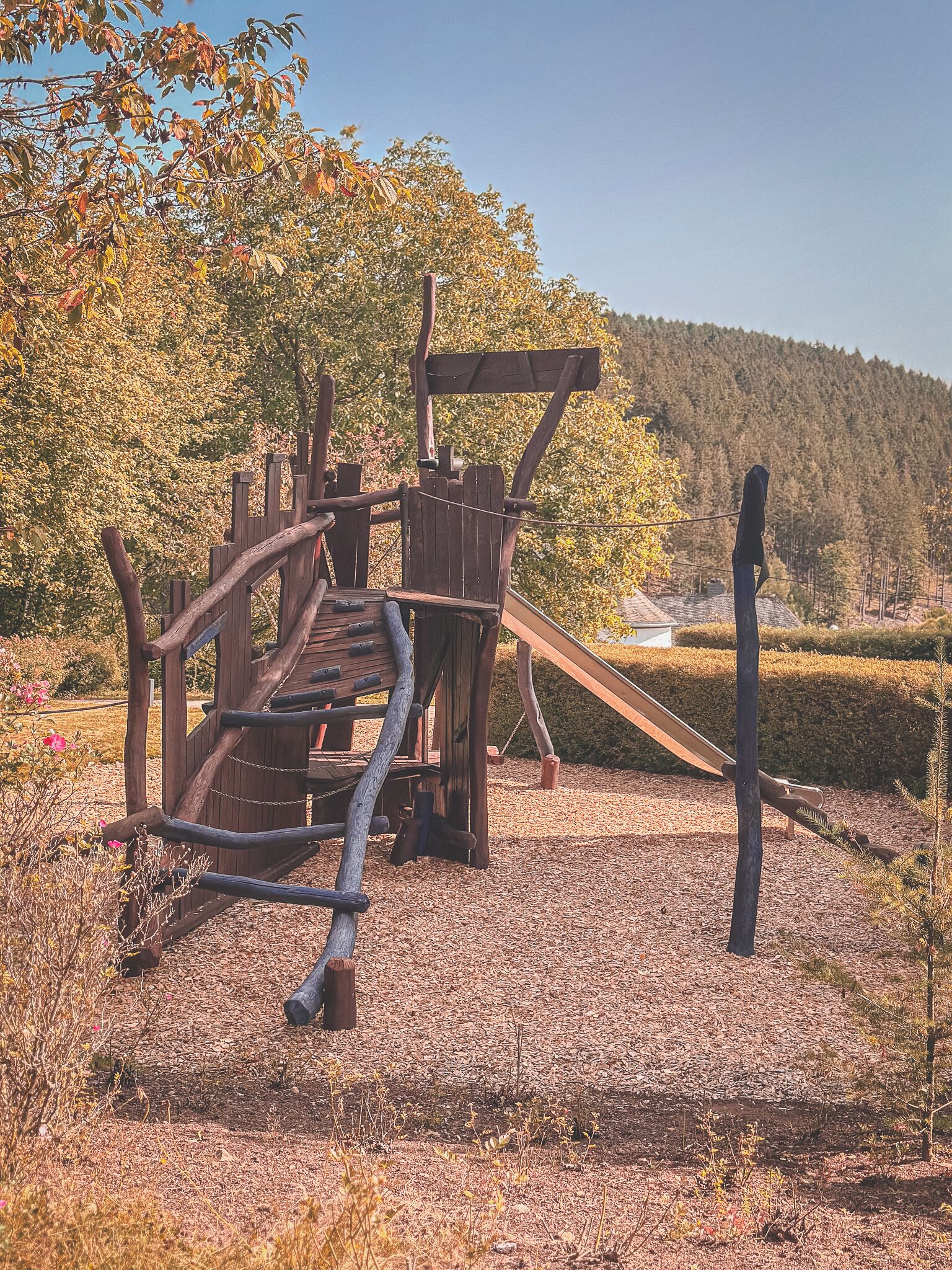 A wooden play structure with slides and climbing elements, surrounded by trees and a clear sky.