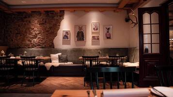 Ein gemütliches Restaurant mit Steinwänden, Holzbalken und einem Kamin aus Stein. Es sind Tische und Stühle zum Essen bereitgestellt, und an den Wänden befinden sich Kunstwerke.
