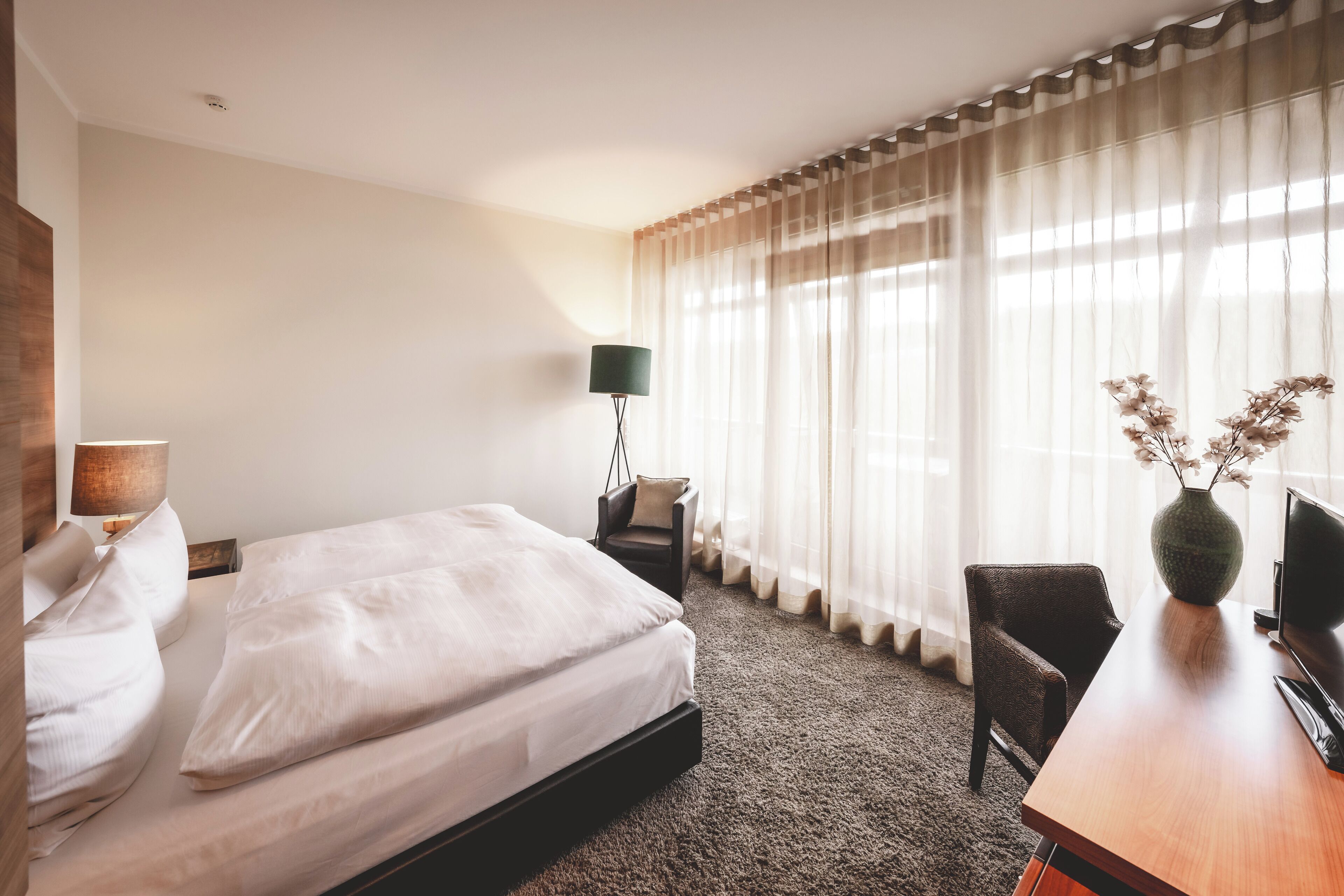 A hotel room with a bed, a desk, a chair, a lamp, a vase with flowers, and sheer curtains.